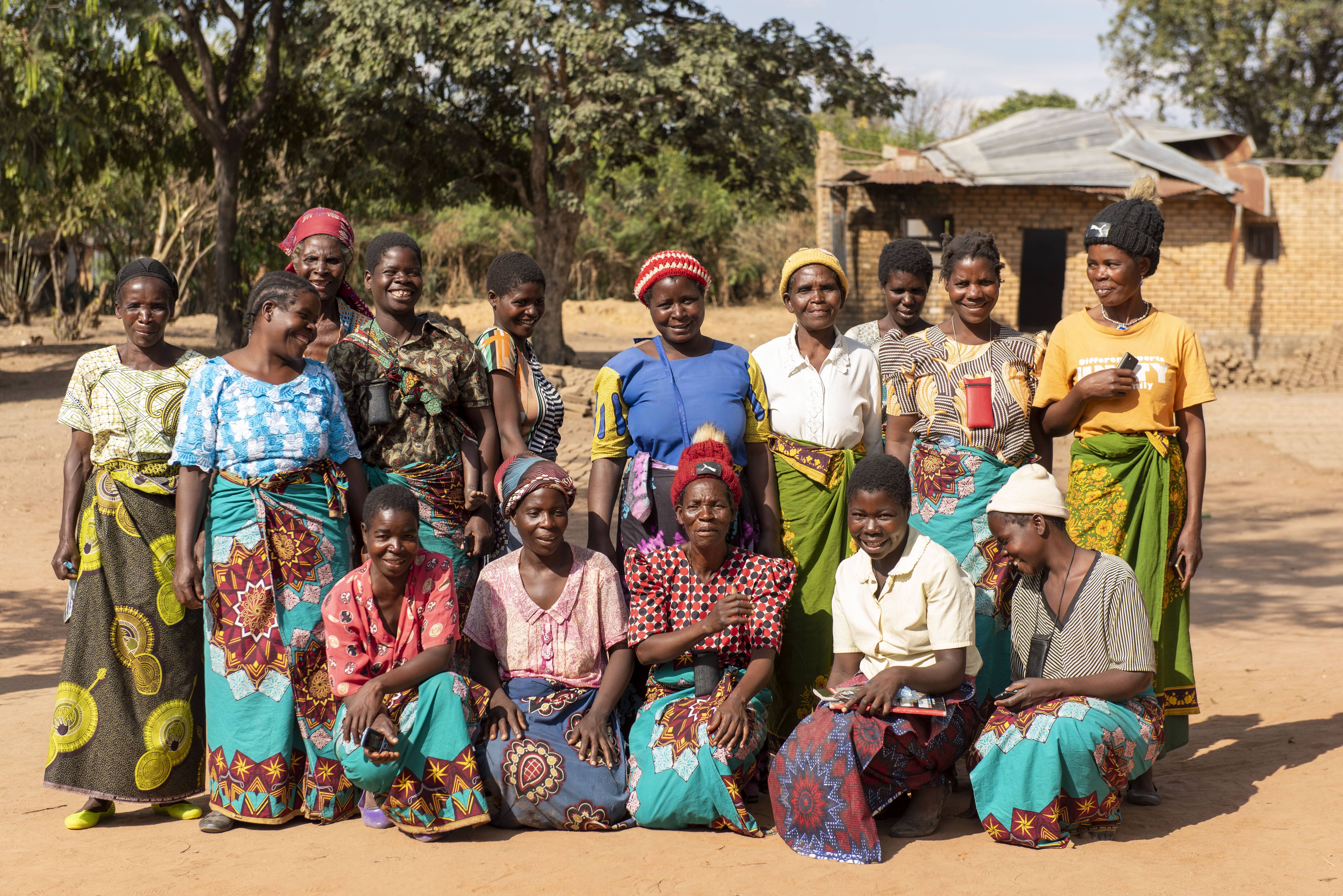 A group of 14 Malawian women stand in 2 rows facing the camera. They're wearing colorful dresses, and most of them are smiling. 
